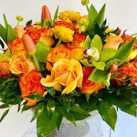 Bouquet of orange and yellow roses with orchids and tulips with greenery in a footed bowl or light coloured container