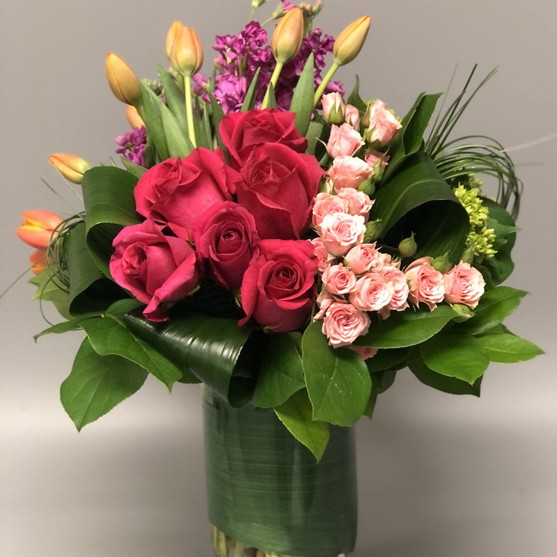 Fuschia Roses, pink spray roses, orange tulips and purple stock with greens designed in clear vase.