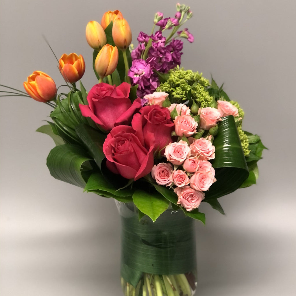 Fuschia Roses, pink spray roses, orange tulips and purple stock with greens designed in clear vase.